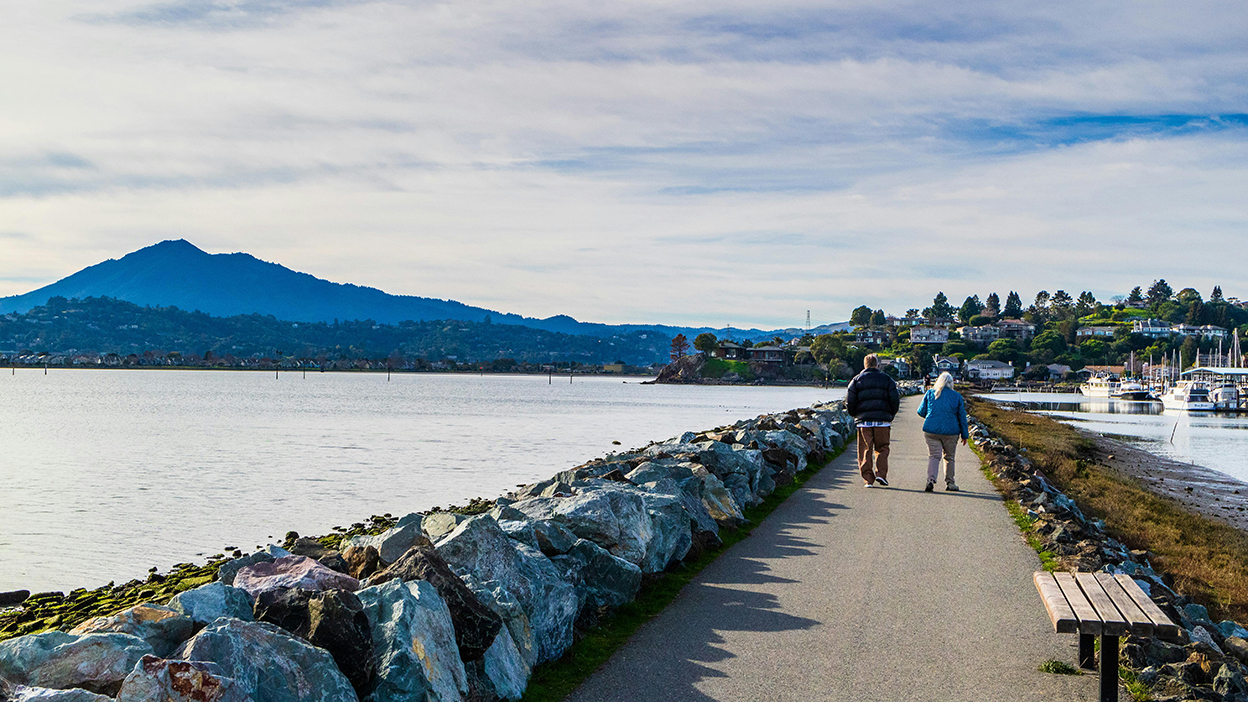 Two people walk down a lane by a lake with a mountain in the background